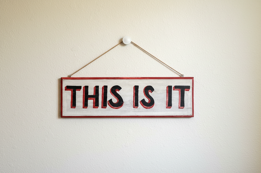 Hand-painted wooden sign reading THIS IS IT hanging on a neutral wall with natural jute rope, featuring red 3D shadow typography on a distressed white background.