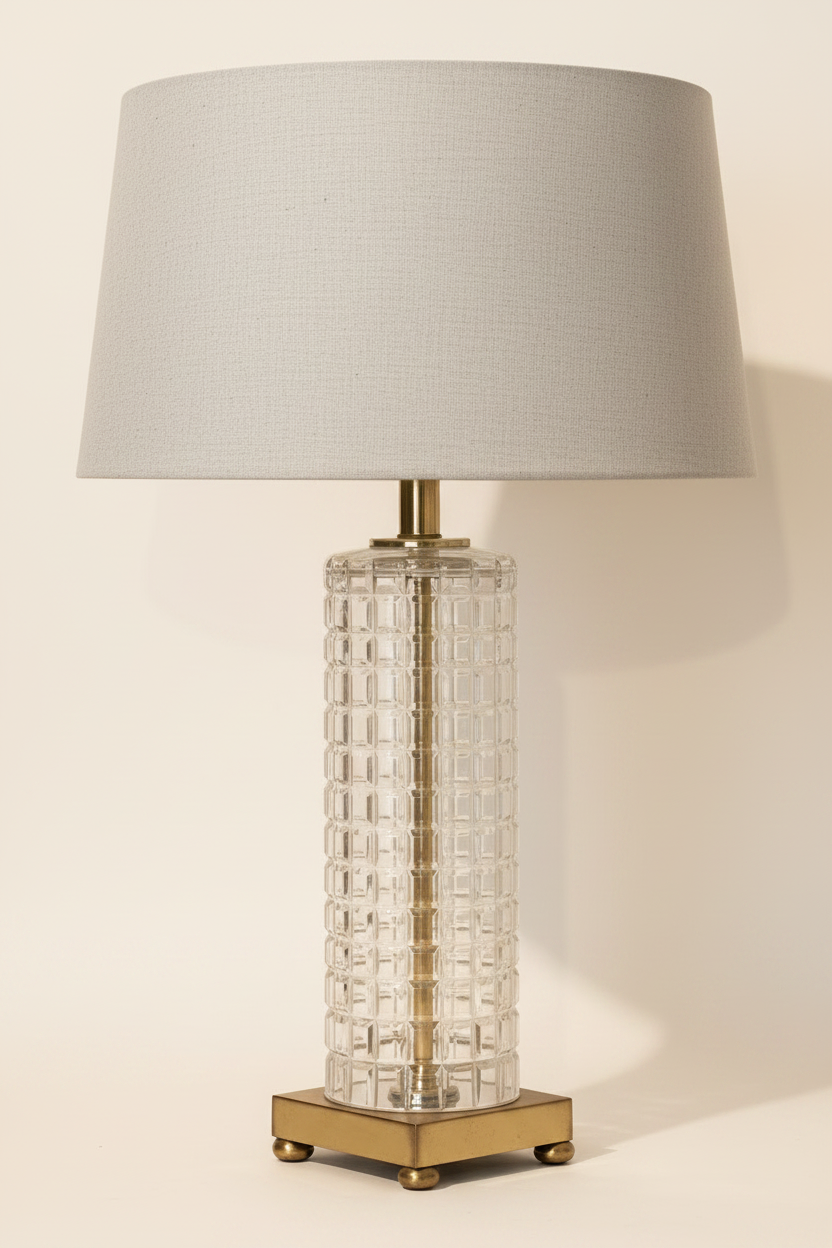 Vintage 1960s 1970s clear pressed glass waffle-grid column table lamp with natural linen drum shade, antique brass stem, and square brass plinth with four ball feet, white studio background, shade not included