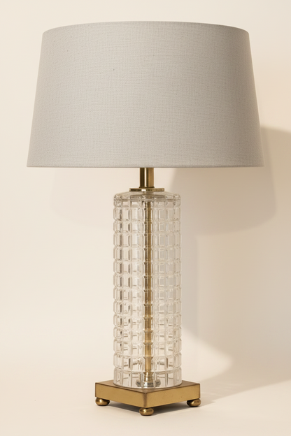 Vintage 1960s 1970s clear pressed glass waffle-grid column table lamp with natural linen drum shade, antique brass stem, and square brass plinth with four ball feet, white studio background, shade not included