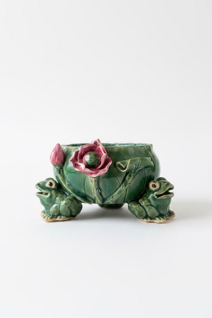 Front view of a vintage mid century ceramic tripod planter with three green figural frogs holding up a leaf basin decorated with a 3D pink water lily flower.