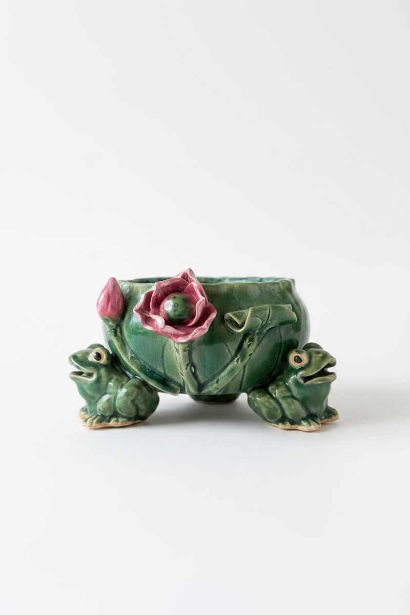 Front view of a vintage mid century ceramic tripod planter with three green figural frogs holding up a leaf basin decorated with a 3D pink water lily flower.