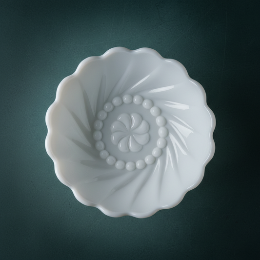 Overhead view inside vintage Fenton white milk glass bowl showing diagonal spiral swirl ribs sweeping inward from scalloped rim to center, raised beaded ring detail surrounding molded 8-petal pinwheel rosette base motif, opaque gloss white pressed milk glass, deep teal background