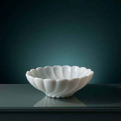 Three-quarter profile of vintage Fenton white milk glass scalloped bowl on dark grey-green marble surface, opaque gloss white pressed glass with diagonal spiral swirl rib pattern and scalloped rim, soft reflection in polished surface, deep teal background