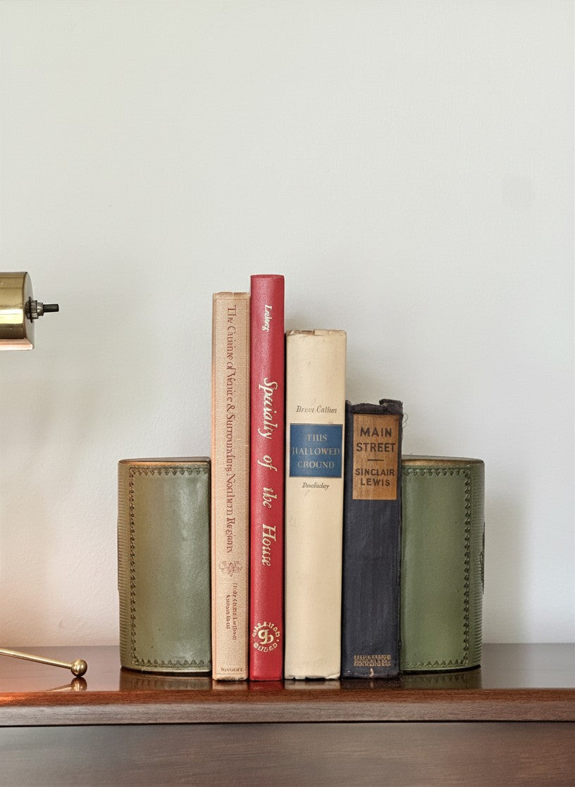 Front view of a pair of vintage 1950s green leather semicircular bookends holding four vintage cloth-bound library books upright on a rich mahogany shelf, styled with a brass desk lamp.